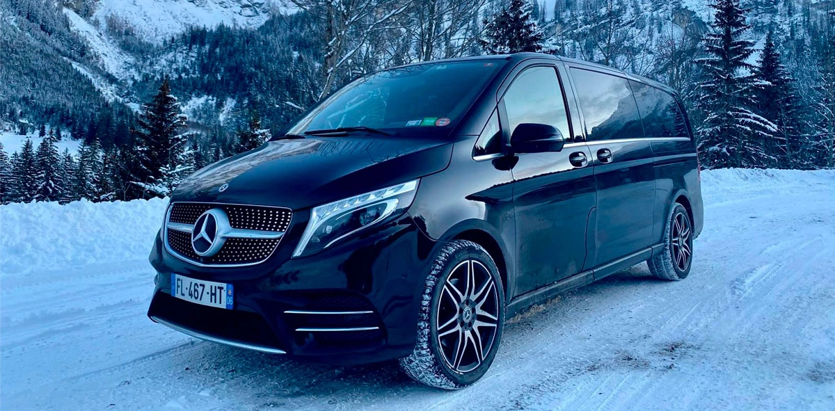 A stylish black Mercedes V-Class van parked on a snow-covered road in a mountainous winter landscape. Surrounded by snow-laden trees and alpine peaks, the vehicle stands out with its sleek design and premium features.