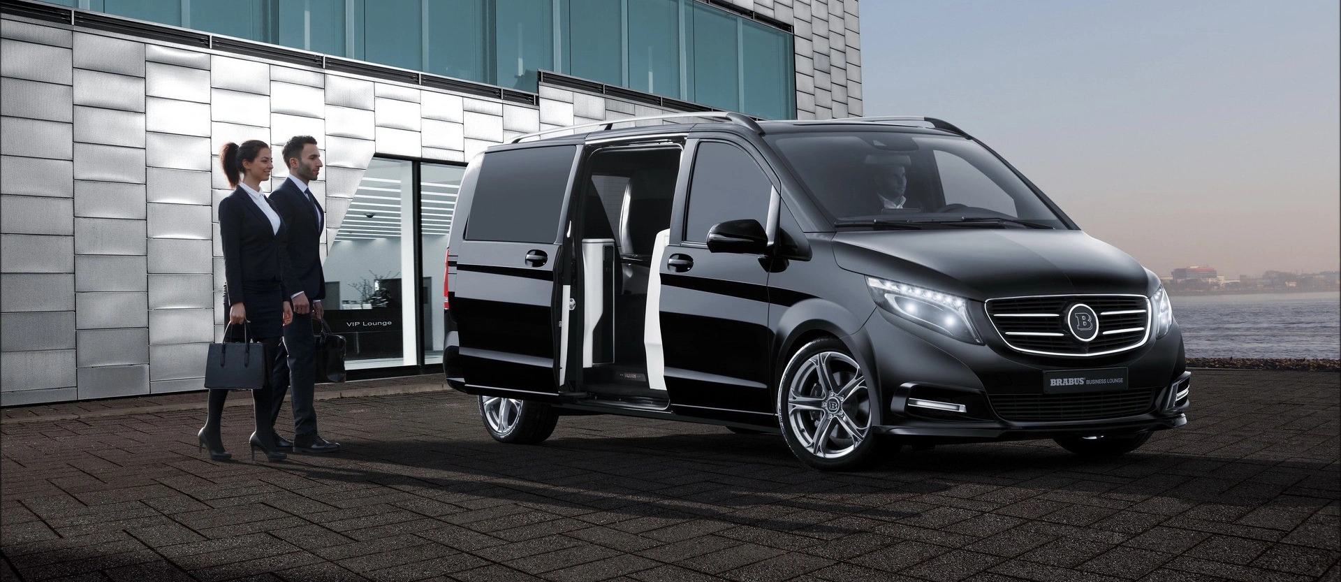 Mercedes V-class parked outside a modern glass building, with two business professionals approaching. The van’s sliding door is open, revealing a premium interior designed for executive travel.