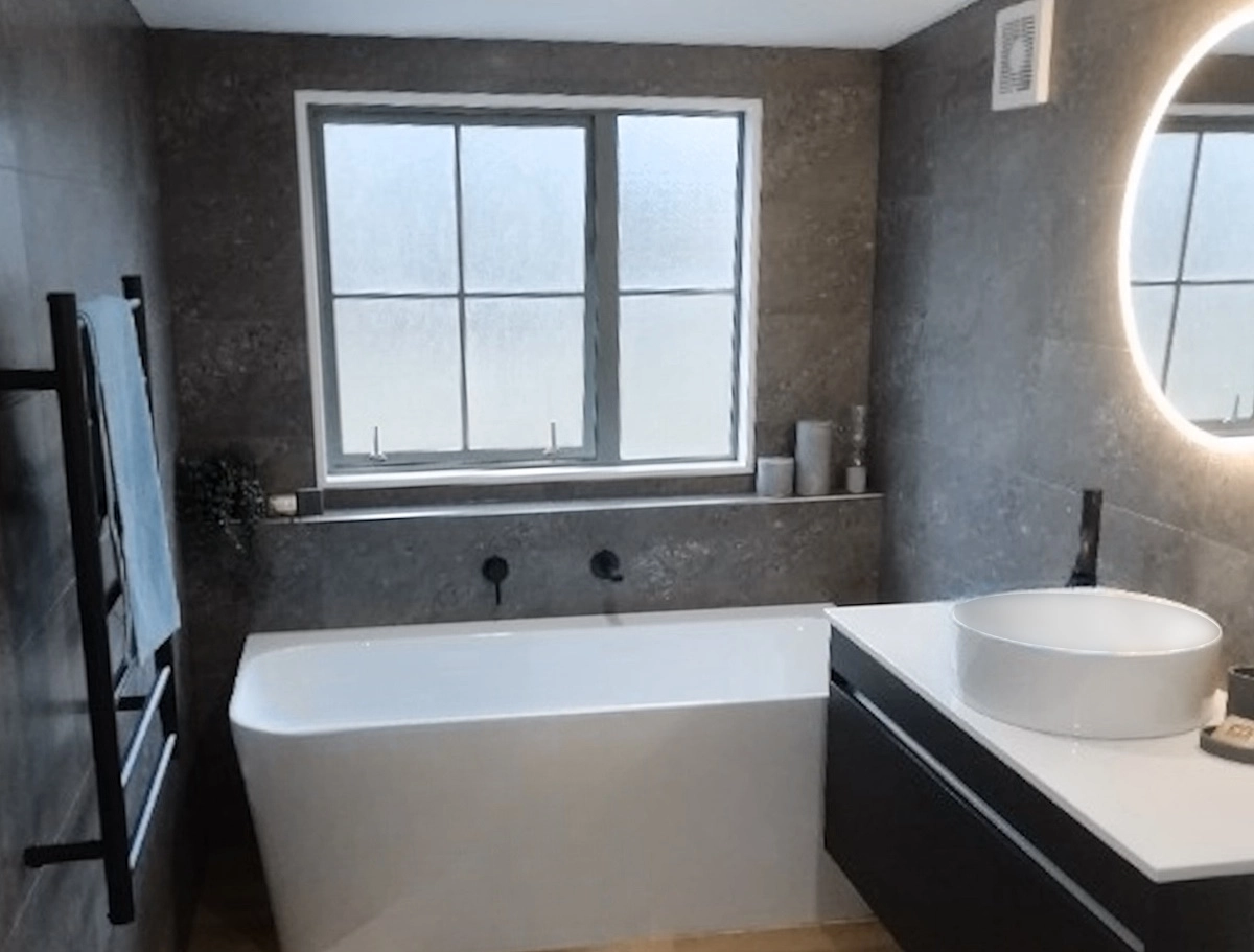 Elegant tiled bathroom in Manor Park featuring dark stone-look tiles, custom shelf behind the bath, and wall-hung vanity – Direct Tiling Wellington