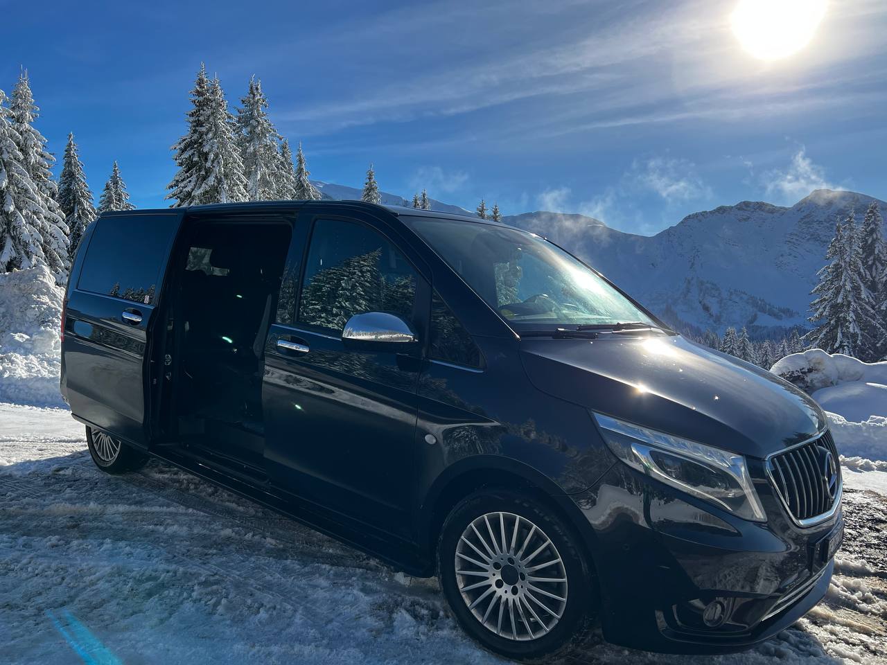 A luxurious black Mercedes V-Class parked on a snow-covered road in a stunning alpine setting. The van's door is open, revealing a premium interior, while the background features snow-laden trees, majestic mountains, and a bright winter sun.