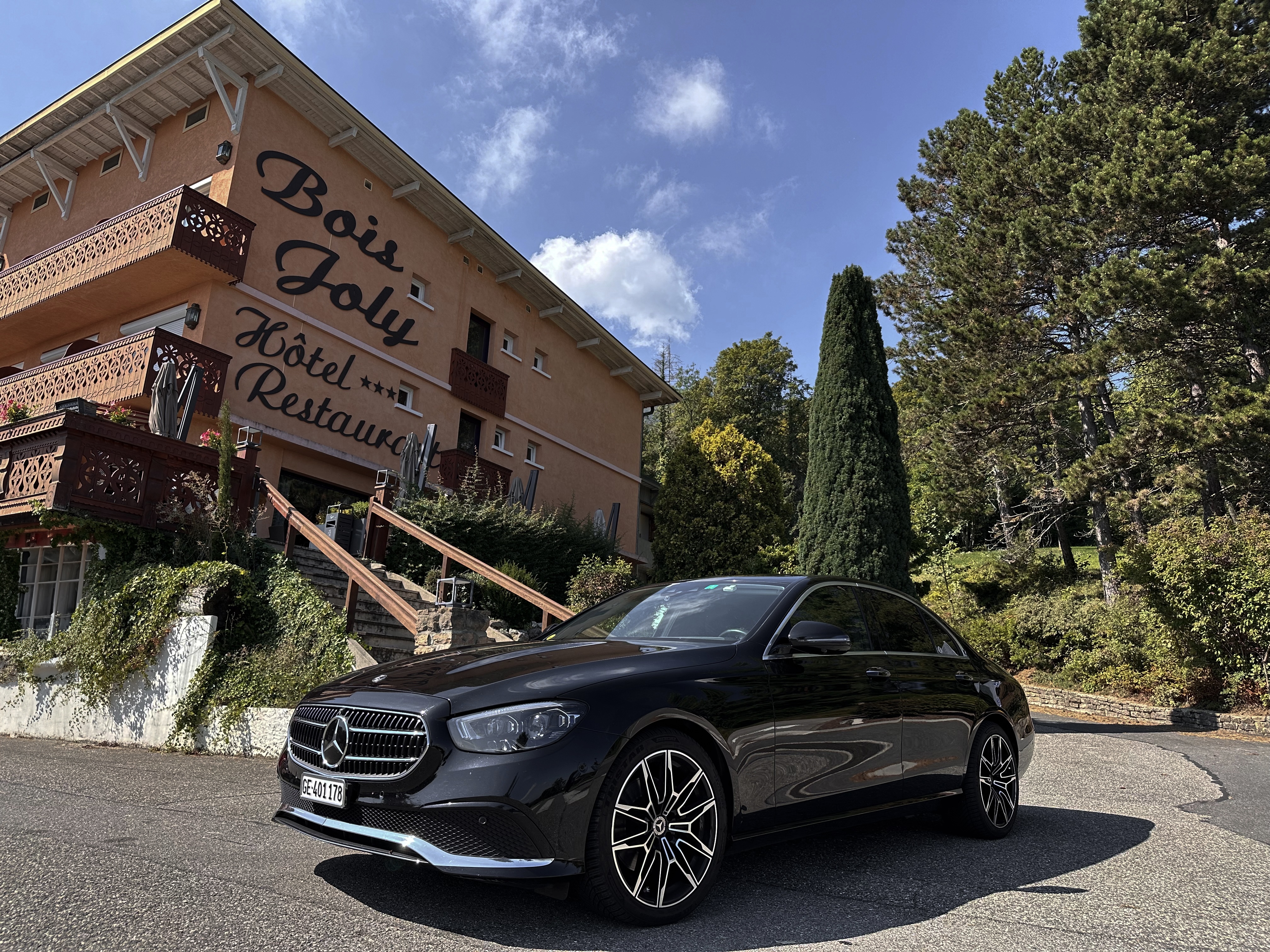 A sleek black Mercedes E-Class TAXI parked in front of the Bois Joly Hotel and Restaurant, a charming building with rustic wooden balconies, surrounded by lush greenery and trees under a sunny blue sky.